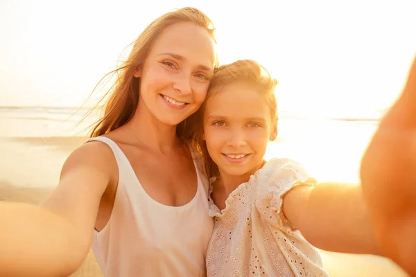 Happy stylishly mother and daughter taking selfie at sandy beach on a sunset.mothers Day.little girl blonde and beautiful woman taking pictures on the phone tourism abroad online video calling