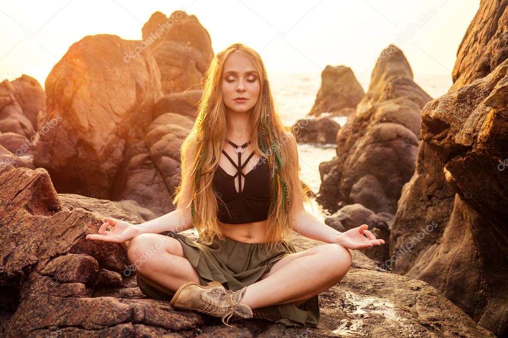 stylish redheaded chestnut blonde young girl in indie bohemian bo-ho style clothes black stylish top.boho woman long hair,makeup and green feathers in head practicing yoga asanas on the rocks by sea