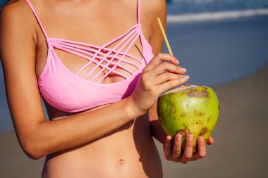 Beyaz kumlu plajda poz hindistan cevizi ile pembe kırmızı mayo güneş gözlüğü ile seksi tabaklanmış model. Mükemmel vücutlu kız yaz gününde tropikal adada oturur. Konsept seyahat, mayo ve detoks