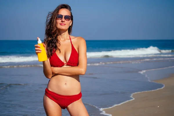 美しい日焼けした女性フィットネスビッグおっぱいモデルは、海のビーチの青空とヤシの木の背景にファッションスタイリッシュな水着で胸に適用spfボトルを保持します。 — ストック写真