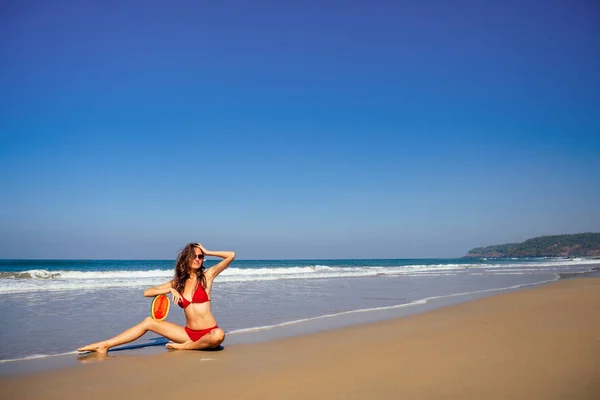 Schöne junge Frau in Badeanzug und roter Sonnenbrille hält Scheibe Wassermelone rot, während sie am Strand Detox steht — Stockfoto