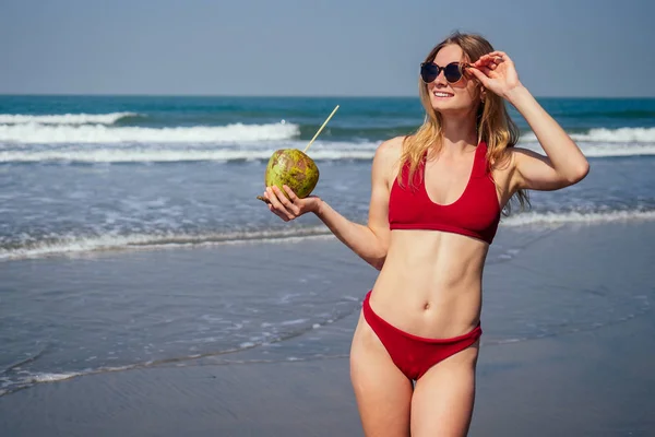 Sexy und junge Frau in einem stylischen Bikini-Badeanzug mit einer Kokosnuss an der Küste des tropischen Paradieses Ozean. sexy perfekte Passform Modell in Wasser mit Kokosmilch Tropfen gießen Kokosnusswasser — Stockfoto