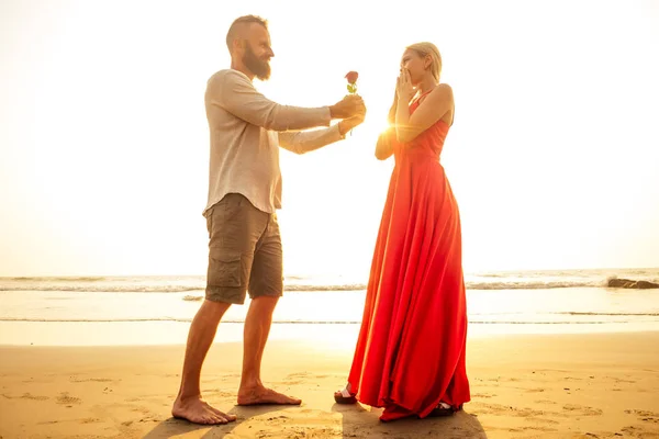 Mann in weißem Hemd überreicht der überraschten Wunderfrau in einem langen roten Kleid am Strand des Meeres Rosen. romantisches Date, Hochzeit oder Valentinstag Konzept von sea.loving Paar feiert 8. März Frauentag — Stockfoto