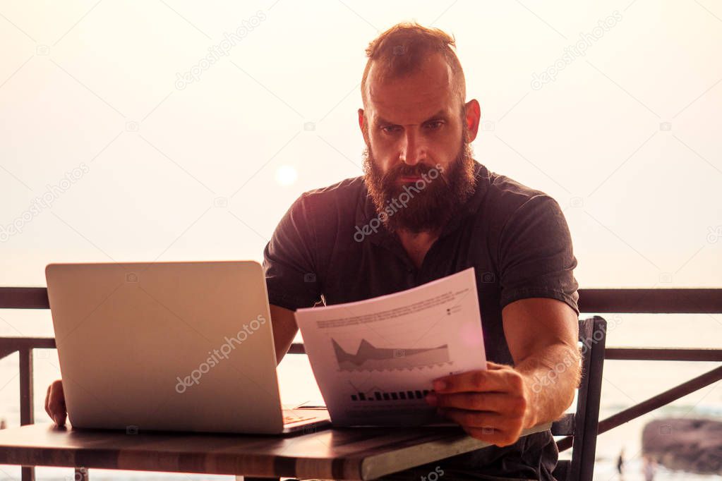 handsome and successful man in a stylish well-dressed freelancer working with a laptop on the beach.freelance and remote work.businessman student in a summer cafe on the shore of india ocean