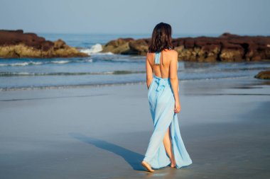 Güzel kadın mavi gökyüzü ve Goa denizi üzerinde bir uçurumun üzerinde duruyor. Esmer kız mavi ipek elbise copyspace güneş kremi spf arka