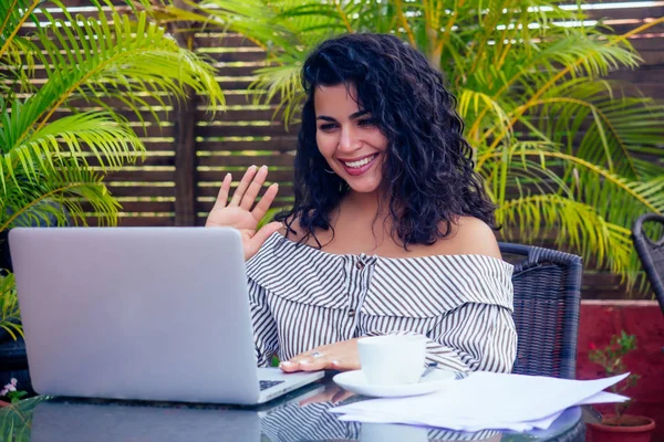 Молодая темнокожая деловая женщина сидит в кафе с ноутбуком, стоящим на столе возле чашки кофе. Фрилансер, работающая индианка, занимающаяся йогой асаной в тропическом курорте — стоковое фото