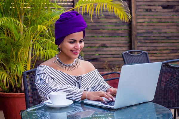 Улыбающаяся красивая индийская девушка с этническим тюрбаном на голове культуры работает на ноутбуке на открытом воздухе летнее кафе — стоковое фото