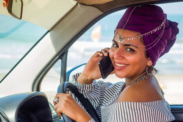 African american caucasian woman in a purple turban head posing with her new car showing the key outdoors, outside in summer day beach ocran sea on background.indian girl ride travel