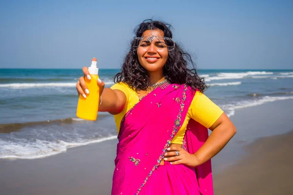 Портрет красивой улыбающейся белоснежной улыбки индианки с черными вьющимися волосами и темной кожей в розовом сари с бутылкой солнцезащитного спрея на пляже. — стоковое фото