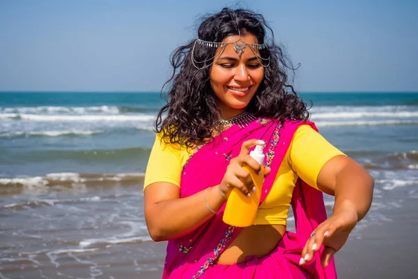 Porträtt av en vacker leende snövit leende indian kvinna svart lockigt hår och mörk hud i en rosa sari hålla flaska solskyddsspray på stranden.flicka njuter spf kropp paradis semester — Stockfoto