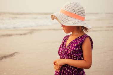 Mor bir elbise ve şapka ve güneş gözlüğü deniz, yaz, tatil, seyahat konsepti yakınındaki plajda rahatlatıcı Pretty küçük bebek kız. plaj tatil spf ve güneş kremi gülümseyen sevimli küçük kız