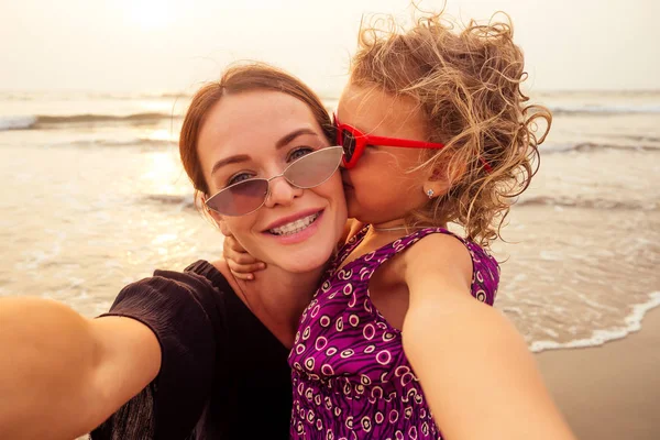 Happy stylishly mother and daughter taking selfie at sandy beach on a sunset.mothers Day.little girl blonde and beautiful woman taking pictures on the phone tourism abroad online video calling