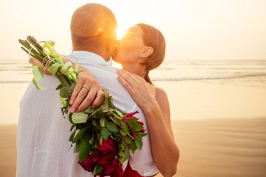 wife and husband happy couple on vacation by the sea.beautiful woman in red dress with a ring on her finger and loving man in a white shirt with a bouquet of roses romantic sunset.Valentines day date clipart