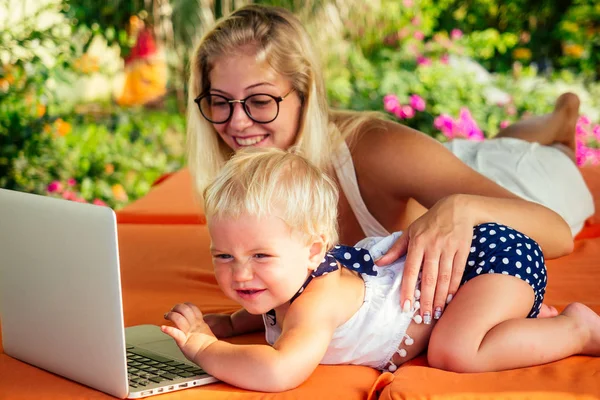 Cabellera de un año de edad, niña mirando a un ordenador portátil de dibujos animados con la madre rubia freelancer surfista en tumbonas resort verano beach.business mujer freelancing y la hija relajarse junto al mar — Foto de Stock