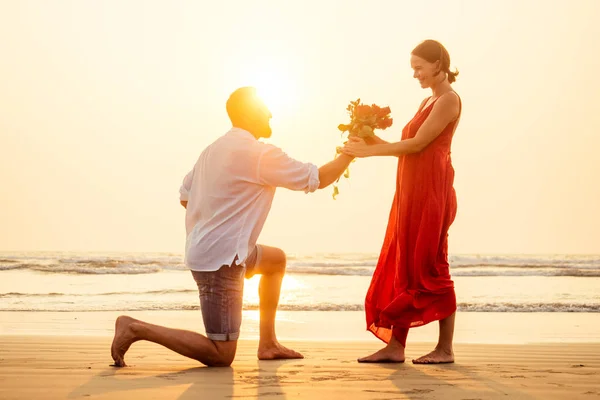 Żona i mąż szczęśliwa para na wakacjach nad morzem. piękna kobieta w czerwonej sukni z pierścieniem na jej palec i kochający człowiek w białej koszulce z bukietem róż romantyczny zachód słońca. Walentynki Data — Zdjęcie stockowe