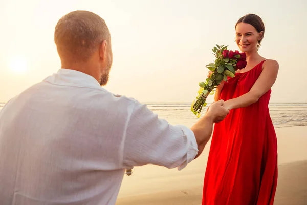 wife and husband happy couple on vacation by the sea.beautiful woman in red dress with a ring on her finger and loving man in a white shirt with a bouquet of roses romantic sunset.Valentines day date