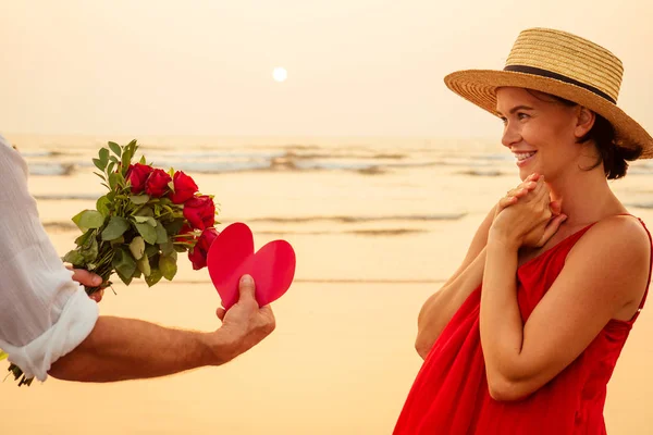 Щаслива красива жінка в червоній сукні отримує подарунок від чоловіка на заході сонця. сюрприз дружина від чоловіка букет троянд жіночий день 8 березня та День святого Валентина. — стокове фото