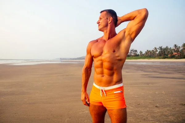 Νέος όμορφος άνθρωπος εφαρμόζοντας κρέμα αντηλιακή λοσιόν στην παραλία της θάλασσας. Σέξι αρσενικό μοντέλο αθλητής bodybuilder θέτοντας το τέλειο αθλητικό σώμα, αποτρίχωση και αίσθηση shugaring — Φωτογραφία Αρχείου