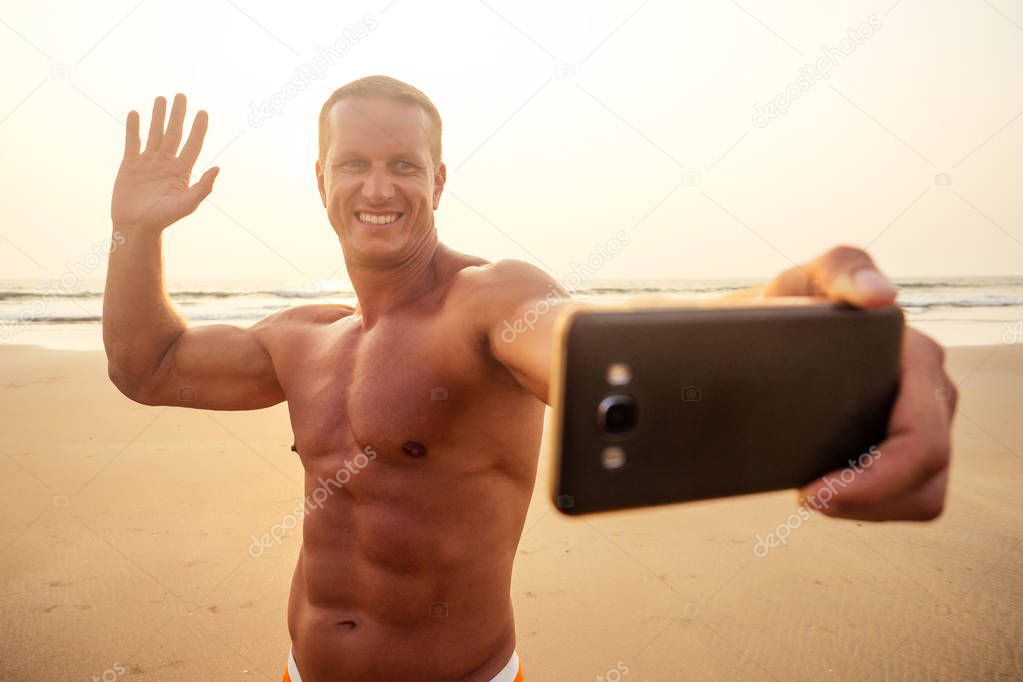 handsome successful male athlete fitness model photographing selfie portrait on phone beach paradise sunset. man on vacation in the tropic talking on the smartphone online with friends and family