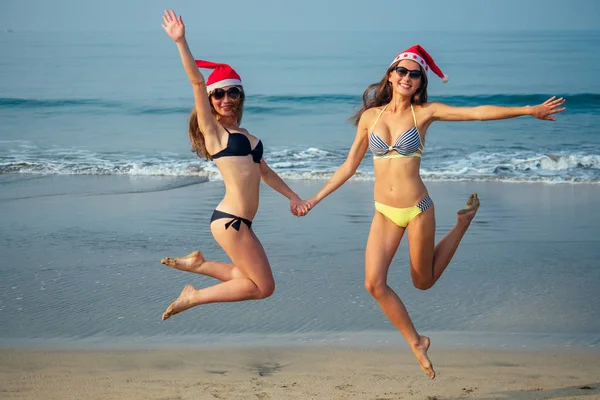 Fröhliche Freundinnen mit Weihnachtsmütze und Bikini springen am Strand — Stockfoto