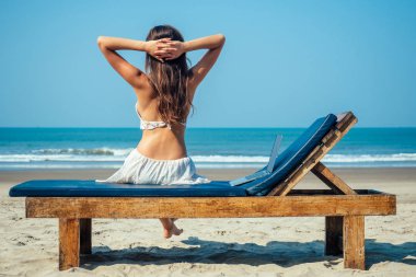 Lotus pozu ve deniz kenarında dizüstü bilgisayarı olan bir şezlongda oturan güzel ve seksi genç bir kadın. Serbest çalışan bir kadın tatil köyünde dizüstü bilgisayarla yoga yapıyor.