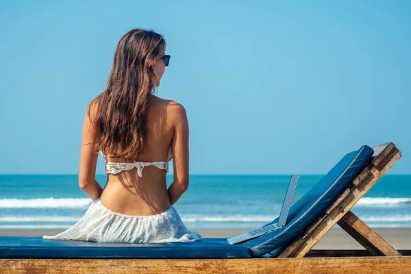 Schöne und sexy junge Frau sitzt in Lotus-Pose und medetiruet auf einer Liege mit einem Laptop am Meer. Freiberuflerin macht Yoga mit Laptop im Resort — Stockfoto