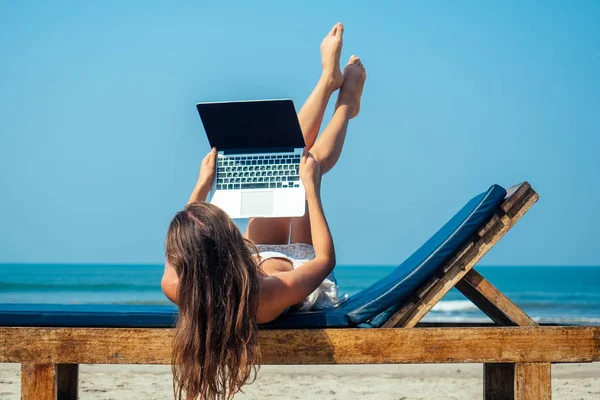 Güzel ve seksi genç bir kadın deniz laptopuyla güverte sandalyesinde uzanıyor ve dinleniyor. Otelde dizüstü bilgisayarı olan serbest çalışan bir kadın.