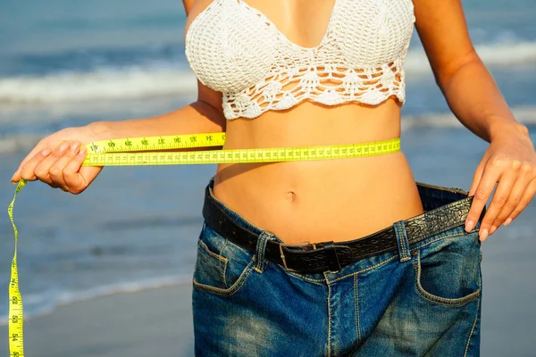 Een jong en slank meisje in spijkerbroek houdt een meetlint in haar hand op haar sexy dijen op het strand. concept van detox en dieet — Stockfoto