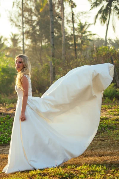 Noiva jovem e bonita com longos cabelos loiros em um vestido de noiva longo branco bonito sorrindo ao fundo das palmeiras. uma noiva feliz se casou com uma cerimônia de casamento na praia junto ao mar — Fotografia de Stock