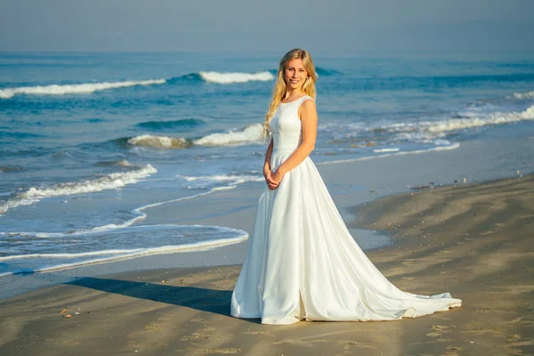 아름답게 웃으며 아름답게 하얀 긴 웨딩 드레스를 입고 바다를 걷고 있는 아름답게 긴 금발의 젊고 아름다운 신부. 해변에서 결혼식을 올리는 행복 한 신부 — 스톡 사진