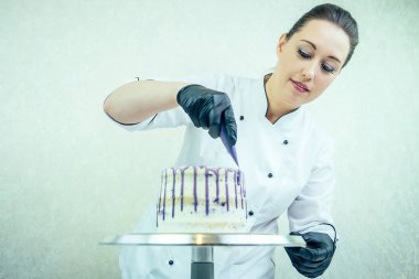 Siyah eldivenler ve beyaz iş üniforması içinde güzel ve gülümseyen kadın pastacı mutfaktaki pastayı süslüyor. Şekerleme, pasta, yemek. düğün ve doğum günü pastası
