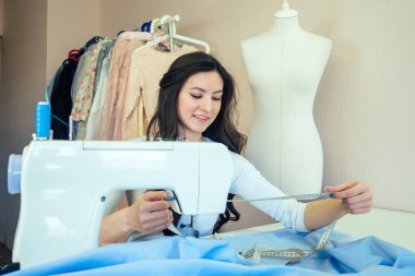 Dikiş makinesiyle çalışan uzun saçlı güzel bir terzinin portresi. Terzi bir kıyafet koleksiyonu yaratıyor. Genç bayan moda tasarımcısı elbise dikiyor.