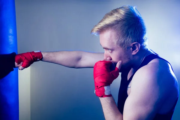A young male boxer is doing sports in the gym. boxer, puts on boxing gloves on a dark background. the man strikes. red bandage on the hands