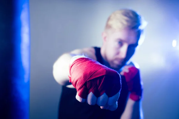 A young male boxer is doing sports in the gym. boxer, puts on boxing gloves on a dark background. the man strikes. red bandage on the hands