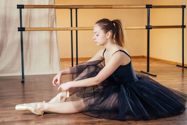 Eine junge, schöne Frau im Ballettkleid schnallt die Spitzenschuhe an ihre Füße. Die Ballerina wärmt sich im Klassenzimmer auf. das Ballerinas-Mädchen in einem schönen schwarzen Kleid. — Stockfoto