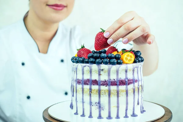 beautiful and smiling woman confectioner in white work uniform adorns the cake in the kitchen. confectioner, cake, cooking. wedding and birthday cake with berries