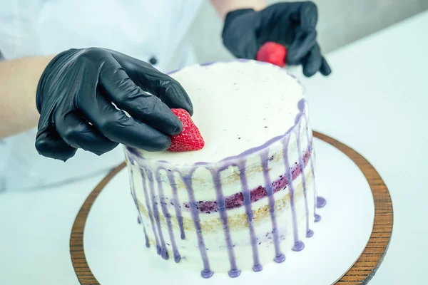 confectioner in black gloves and white work uniform adorns the cake in the kitchen. confectioner, cake, cooking. wedding and birthday cake with berries