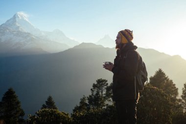 Dağlarda kızıl sakallı ve sırt çantalı genç bir adam elinde sıcak bir içecekle bir fincan tutuyor. Dağlarda aktif eğlence ve turizm kavramı. Baharda Nepal
