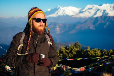 Dağlarda kızıl sakallı ve sırt çantalı genç bir adam elinde sıcak bir içecekle bir fincan tutuyor. Dağlarda aktif eğlence ve turizm kavramı. Baharda Nepal