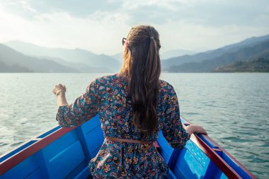 Güzel elbiseli genç kadın turist tekneyle gölde yüzüyor ve dağların manzarasının tadını çıkarıyor. Açık hava aktiviteleri konsepti. arka plan