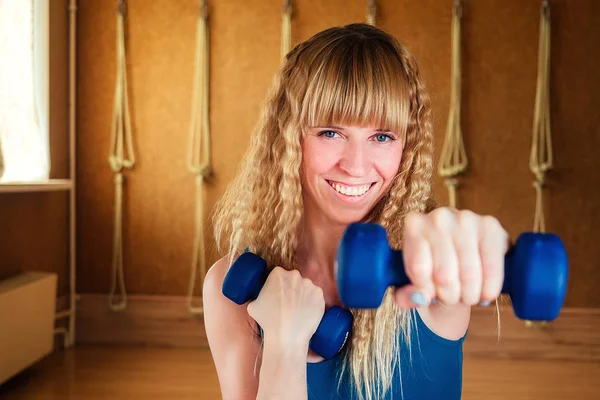 young and beautiful blond woman is holding dumbbells in the hands of a gym. The yoga instructor performs respiratory practice and strengthens the muscles of the hands.