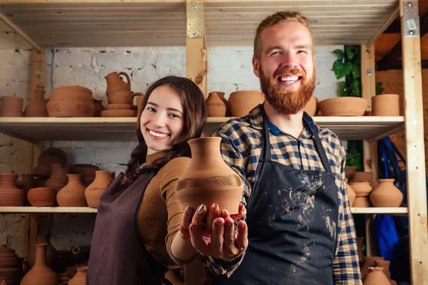 a bearded man and a young woman holding a vase of clay and having fun near the shelf with vases and pots of clay. potter, clay, vase, pottery workshop. master and student