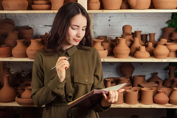 beautiful woman writes in a notebook in a pottery workshop. The potter writes down ideas on a background of a rack with clay pots and vases. Inspiration and Muse