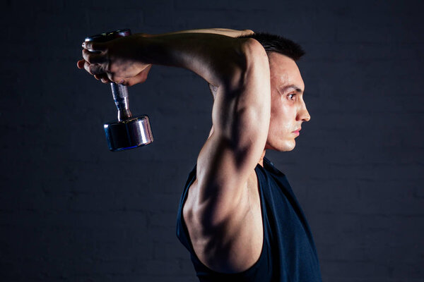 young man strengthens the muscles of the hands of dumbbells in the gym on a dark background