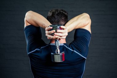 Spor salonunda siyah tişörtlü yakışıklı bir adam ve arkasında bir dambıl var.