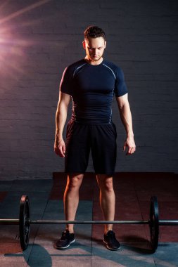 Siyah tişörtlü genç ve yakışıklı bir adam siyah tuğlalı arka planda barın yanında duruyor.