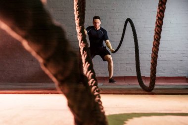 Siyah tişörtlü genç ve yakışıklı bir adam. Salonda iplerle bağlanmış. Duvarın arkasında.