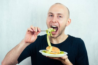 Elinde mezura olan genç ve kel bir adamın portresi yüksek kalorili büyük bir burgeri çatalla bir tabakta yiyor. Diyet kavramı, sağlıksız ve sağlıklı beslenme