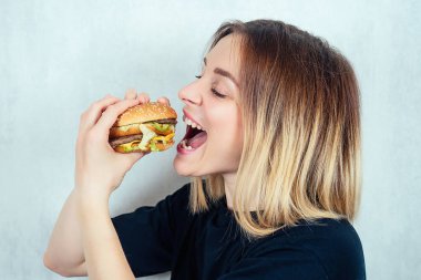 Siyah tişörtlü ve yüksek kalorili burger yiyen genç ve çekici sarışın kadın. Zararlı fast food ve diyet kavramı