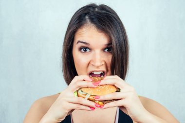 Siyah tişörtlü ve yüksek kalorili burger yiyen genç ve çekici bir kadın.